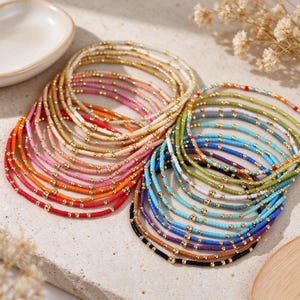 Puede incluir: Una colección de pulseras de cuentas multicolores con pequeños espaciadores redondos de metal dorado intercalados entre cuentas circulares de acabado mate en tonos vibrantes de rojo, naranja, rosa, amarillo, verde, azul, morado y negro.