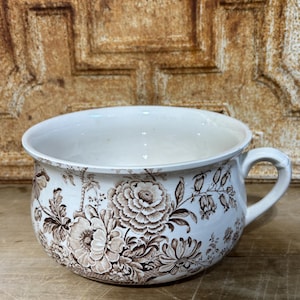 May include: A white ceramic chamber pot featuring a brown floral transferware pattern. The vessel has a wide, circular opening, a curved handle on the right side, and a rounded body decorated with detailed illustrations of various blooming flowers.