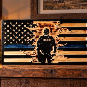 May include: A rectangular wooden wall art piece featuring a carved American flag design with a distressed, burnt-wood finish. A silhouette of a sheriff stands in the center, and a thin blue line runs horizontally across the flag's stripes.