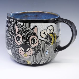 May include: Ceramic mug with a black handle and rim, featuring a whimsical design. The mug showcases a black and white squirrel with pink accents, a yellow and black bee, and blue raindrops. The interior is a vibrant blue.