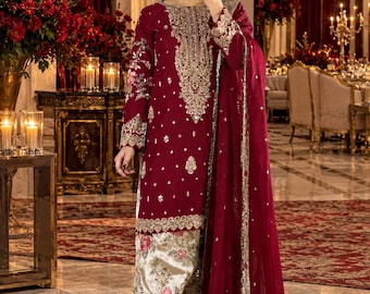 May include: A deep maroon long-sleeved tunic featuring intricate gold embroidery around the neckline and cuffs, paired with cream-colored wide-leg trousers adorned with a floral print and a matching maroon sheer dupatta with gold-trimmed borders.