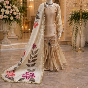 May include: A beige silk-like tunic and flared pants set with gold trim, paired with a cream-colored dupatta featuring a hand-painted floral vine pattern in red, pink, and dark gray, finished with a delicate gold metallic border along the edges.