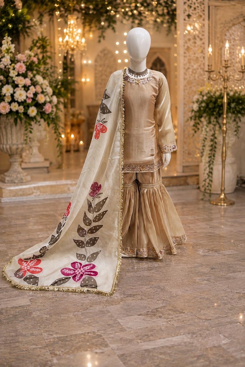 May include: A beige silk-like tunic and flared pants set with gold trim, paired with a cream-colored dupatta featuring a hand-painted floral vine pattern in red, pink, and dark gray, finished with a delicate gold metallic border along the edges.