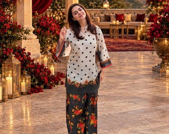 May include: A long-sleeved cream tunic featuring black polka dots and a floral border at the hem, paired with loose-fitting black trousers adorned with a vibrant orange and red floral print. The outfit includes a shoulder bag with a brown strap.