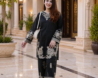 May include: A black long-sleeved tunic and matching wide-leg trousers set featuring intricate cream-colored embroidery along the neckline, sleeve cuffs, and hemline. The outfit is paired with a small white shoulder bag and open-toe slide sandals.