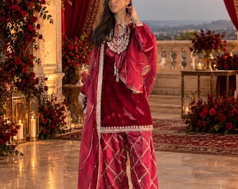 May include: A deep red velvet tunic featuring intricate white embroidery around the neckline and hem, paired with matching wide-leg trousers with a diamond-patterned metallic overlay and a sheer red dupatta with a decorative border.