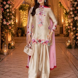 Könnte beinhalten: Eine champagnerfarbene Seidentunika und eine passende Hose mit weitem Bein, verziert mit aufwendigen Blumenstickereien in Rosa, Rot und Blau. Das Outfit umfasst ein hellrosa, transparentes Dupatta und eine kleine, gesteppte beige Handtasche mit goldenen Beschlägen.