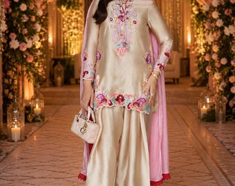 May include: A champagne-colored silk tunic and matching wide-leg trousers featuring intricate floral embroidery in shades of pink, red, and blue. The outfit includes a light pink sheer dupatta and a small, quilted beige handbag with gold hardware.
