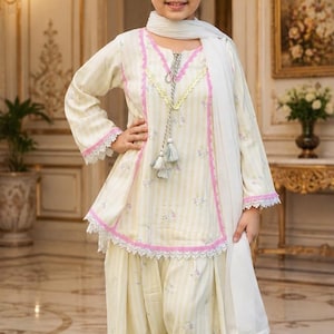 May include: A pale yellow and white striped tunic with pink lace trim, long sleeves, and a white tassel drawstring. The outfit includes matching loose-fitting trousers with a subtle floral pattern and a plain white scarf draped over the shoulders.