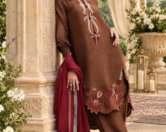 May include: A brown long-sleeved tunic featuring intricate embroidery around the neckline and hem, paired with matching brown trousers and a deep red sheer dupatta. The tunic has a split hem and cuffed sleeves with detailed stitching.