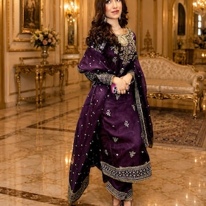 May include: A deep purple long-sleeved tunic and matching trousers set featuring intricate gold embroidery around the neckline, cuffs, and hem. The outfit includes a coordinating sheer purple dupatta draped over the shoulders with scattered gold embellishments.
