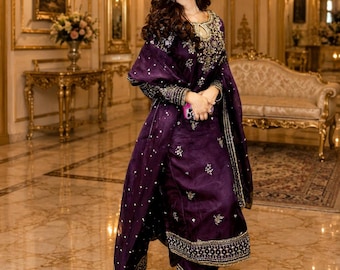 May include: A deep purple long-sleeved tunic and matching trousers set featuring intricate gold embroidery around the neckline, cuffs, and hem. The outfit includes a coordinating sheer purple dupatta draped over the shoulders with scattered gold embellishments.
