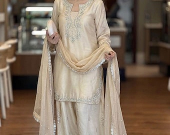 May include: A beige three-piece ensemble consisting of a long-sleeved tunic, wide-leg trousers, and a sheer dupatta. The tunic features a notched neckline with silver embroidery, while the trousers and dupatta are adorned with matching silver floral motifs.