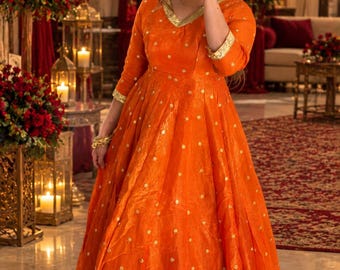 May include: A vibrant orange floor-length dress featuring a V-neckline, three-quarter sleeves, and a flared skirt. The garment is adorned with small gold polka dot patterns and finished with a wide, textured gold metallic border at the hem.