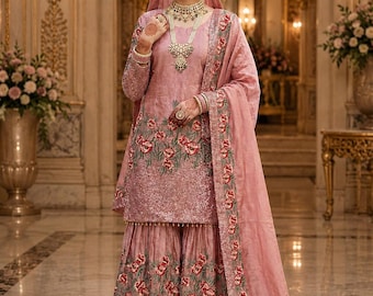 May include: A dusty rose sharara suit featuring a long-sleeved tunic and wide-leg pants with intricate floral embroidery in red and green. The ensemble includes a matching dupatta with a decorative border and heavy sequin embellishments throughout.