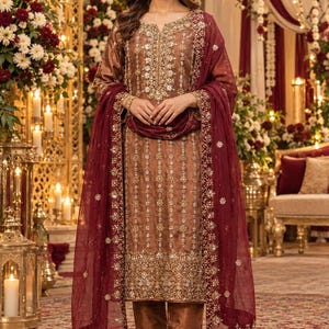 Könnte beinhalten: Ein braunes dreiteiliges Set bestehend aus einer langärmeligen Tunika, einer Hose mit geradem Bein und einem transparenten kastanienbraunen Dupatta. Tunika und Hose sind mit aufwendigen Goldstickereien, Perlen und Pailletten verziert.