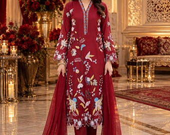 May include: A deep red long-sleeved tunic featuring a V-neckline with blue and white trim, adorned with multi-colored floral embroidery. The garment is paired with matching red trousers and a sheer red dupatta draped over the arms.
