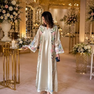 May include: A floor-length champagne-coloured satin kaftan featuring a V-neckline with white beaded trim and long, wide sleeves. The garment is decorated with embroidered floral patterns in shades of pink, blue, and green along the front and sleeves.