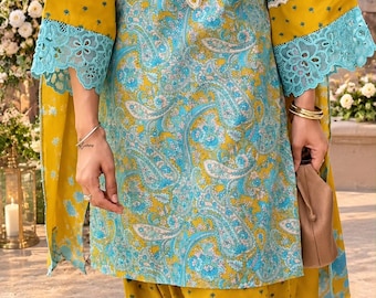 May include: A mustard yellow and light blue tunic set featuring a paisley print. The tunic has a V-neckline with white scalloped trim, three-quarter length sleeves with wide light blue lace cuffs, and matching mustard yellow trousers with a small floral pattern.