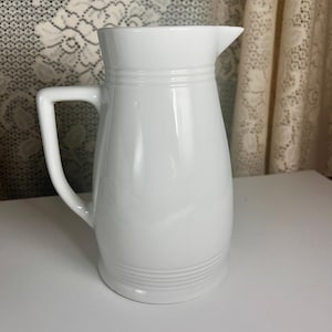 May include: A solid white ceramic pitcher featuring a smooth, glossy finish, a curved handle on the side, a pointed pouring spout, and subtle horizontal ridged detailing encircling the neck and the base of the vessel for a clean, simple look.