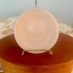May include: A round, pale pink translucent glass plate featuring concentric circular ridges along its outer edge. The plate is held upright by a thin, gold-toned metal wire display stand that features a hook at the top for hanging purposes.