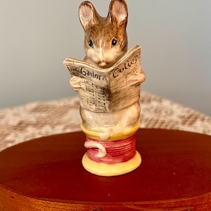 May include: A ceramic figurine of a brown and white mouse sitting on a yellow and red thread spool. The mouse is holding an open newspaper with the text The Tailor & Cutter printed on the front. The mouse has large ears and dark eyes.