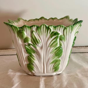 May include: A ceramic planter shaped like a head of lettuce with a cream-colored base and vibrant green painted details on the ruffled, leaf-like edges. The vessel features a wide, open top and a textured exterior mimicking organic foliage.