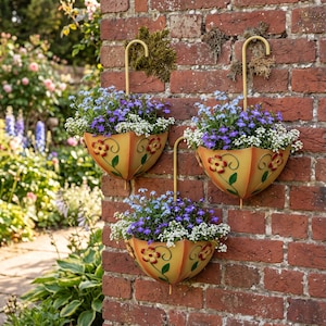 May include: Three yellow umbrella-shaped wall planters with curved handles, each featuring painted red and yellow flowers with green stems. The planters are filled with a mix of small purple and white flowers, mounted on a red brick wall.