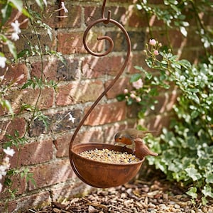May include: A rust-coloured metal hanging bird feeder featuring a curved, scroll-shaped hook and a bowl-shaped basin filled with mixed birdseed. A small, realistic bird figurine is attached to the rim of the bowl, which is suspended in the air.
