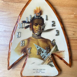 May include: An arrowhead-shaped wall clock with a glossy amber-colored resin border. The clock face features a portrait of Moa-Na-Hon-Ga, an Ioway Chief, with silver-toned metallic numerals and clock hands. Text at the bottom reads: MOA-NA-HON-GA. Great Walker. AN IOWAY CHIEF.