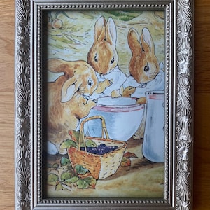 May include: A rectangular silver-toned frame with ornate floral and beaded detailing containing a watercolor illustration of three brown rabbits gathered around a white mixing bowl, a woven basket filled with dark berries, and a white pitcher.