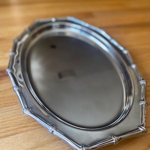 Puede incluir: Una bandeja de servicio ovalada de color plateado con una superficie pulida y reflectante, y un borde elevado con un diseño segmentado inspirado en el bambú. La bandeja tiene un centro liso y plano, y un borde decorativo distintivo.