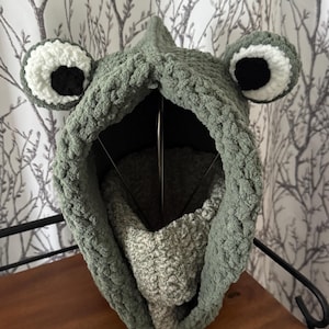 May include: A sage green crochet balaclava featuring a textured, chunky yarn construction with two prominent, rounded frog-like eyes on top. The eyes are white with large black pupils. A lighter, speckled gray-green neck warmer is visible inside.