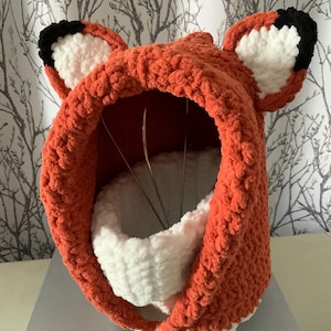 May include: A crochet fox hood made of chunky orange yarn with white and black ear details. The hood features a soft white interior lining and a matching white neck warmer piece visible inside the opening, crafted with a textured, plush yarn finish.