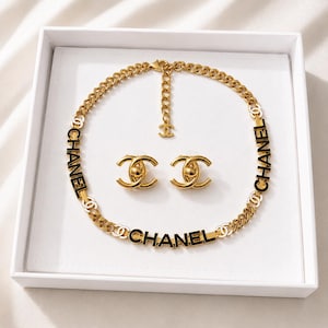 May include: A gold-tone chain link necklace featuring the black text CHANEL integrated into the links, accompanied by a pair of matching gold-tone stud earrings with a central circular element and a dangling CC logo charm on the necklace extender.