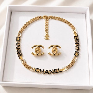 May include: A gold-tone chain link necklace featuring the black text CHANEL integrated into the links, accompanied by a pair of matching gold-tone stud earrings shaped like the interlocking CC logo with a central sphere detail.
