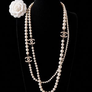 May include: A long multi-strand necklace featuring cream-colored pearls of varying sizes and three gold-toned CC logos encrusted with clear crystals. A white camellia flower brooch is positioned to the left of the necklace strands.