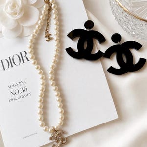 May include: A white pearl necklace featuring a gold-tone CC logo charm and an adjustable chain with a small CC pendant, paired with two large black acrylic CC logo earrings. The jewelry is displayed on a white Dior Magazine cover.