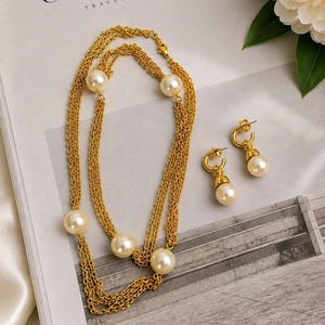 May include: A gold-toned multi-strand chain necklace featuring five evenly spaced cream-colored faux pearls. The set includes a pair of matching drop earrings, each consisting of a small gold-toned hoop with a single cream-colored faux pearl pendant.