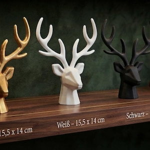 Può includere: Tre sculture di teste di cervo in oro, bianco e nero, ognuna con un design geometrico e corna prominenti. Le etichette testuali indicano dimensioni di 15,5 x 14 cm per ogni pezzo, etichettate come Gold, Weiß e Schwarz.