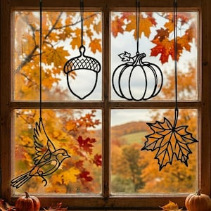 Könnte beinhalten: Vier schwarze Metalldraht-Ornamente hängen in einem Fenster, bestehend aus einer Eichel, einem Kürbis, einem fliegenden Vogel und einem Ahornblatt. Jedes Ornament ist an einer dünnen schwarzen Schnur vor herbstlich gefärbten Bäumen befestigt.