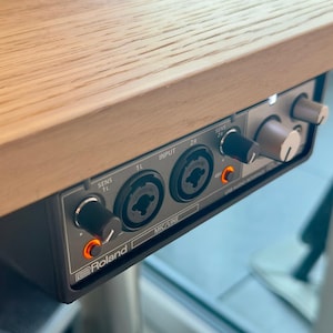 Könnte beinhalten: Ein schwarzes Roland USB-Audio-Interface, das unter einem hellen Holztisch montiert ist. Die Vorderseite verfügt über zwei XLR/TRS-Kombi-Eingänge, zwei Empfindlichkeitsregler, einen großen Lautstärkeregler und zwei orangefarbene 48V-Leuchten.