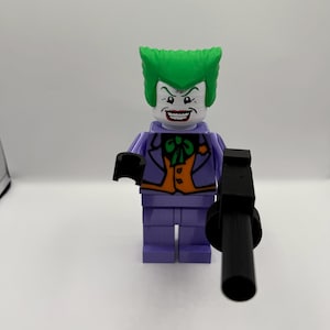 May include: A Joker minifigure with bright green hair, a white face, and a wide red-lipped grin. The figure wears a purple jacket over an orange waistcoat with a green bowtie and purple trousers. It holds a black plastic toy gun in its right hand.