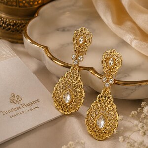 May include: A pair of gold-tone drop earrings featuring intricate filigree metalwork and clear, marquise-cut crystals. Each earring has a teardrop-shaped base and a smaller top section connected by three small round stones. A card reads Timeless Elegance.