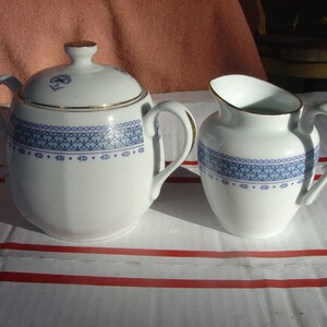 May include: A white porcelain sugar bowl and creamer set with a blue and gold geometric pattern. The sugar bowl has a lid and both pieces have a gold trim.