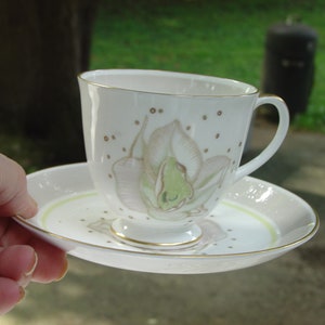 May include: A white teacup and saucer with a gold rim. The teacup has a floral design with a green and white orchid. The saucer has a light green band around the edge.