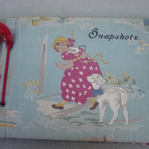 May include: Vintage photo album with a light blue cover featuring a whimsical illustration of a girl in a pink dress with a lamb. The word "Snapshots" is printed on the cover. A red tassel and cord secure the album.