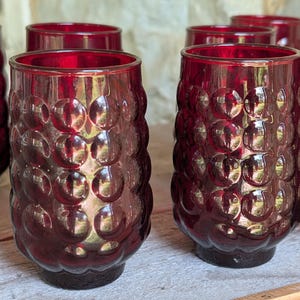 May include: A collection of ruby red drinking glasses. The glasses have a textured, bubble-like pattern on the exterior. The glasses are cylindrical with a slightly flared rim and a dark base. The glasses are arranged on a light-colored wooden surface.