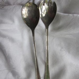 Puede incluir: Un par de utensilios de servir plateados, una cuchara y un tenedor, se muestran sobre un fondo blanco. La cuchara tiene un cuenco redondeado, mientras que el tenedor tiene tres púas. Ambos utensilios tienen mangos largos y delgados con un diseño decorativo de concha.