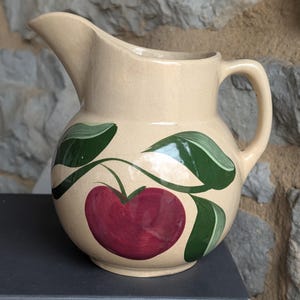 Puede incluir: Jarra de cerámica color crema con cuerpo redondeado, pico y asa. La jarra está decorada con una gran manzana roja y hojas verdes. La jarra se utiliza probablemente para servir bebidas.